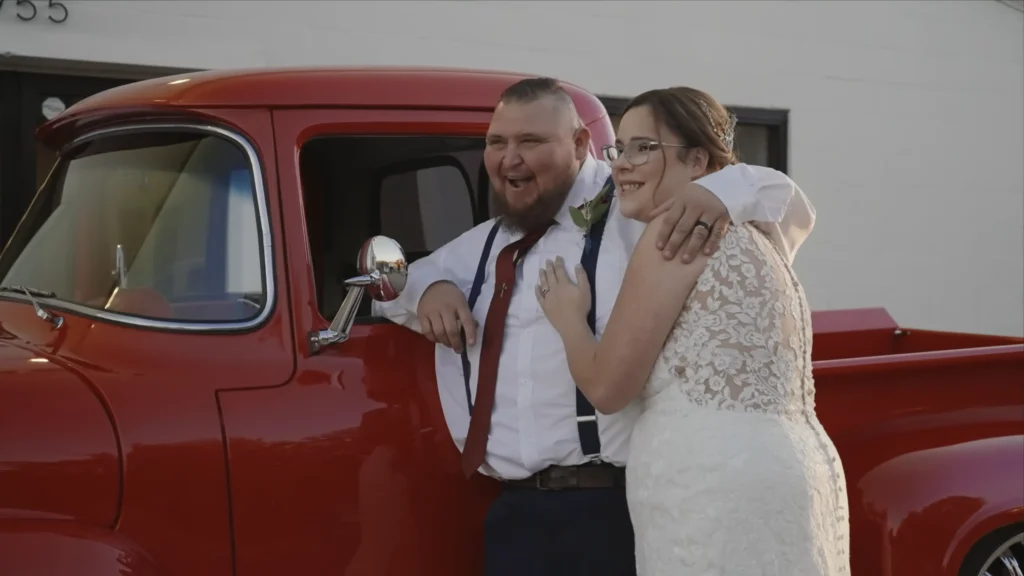 Bride and groom laughing together beside a vintage red truck during their wedding celebration